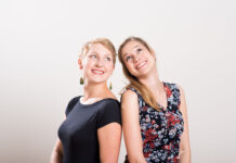 SPRINGTIME! / Kulturforum New York Das Bild zeigt zwei junge Frauen, die lächelnd und mit einem Ausdruck der Freude in die Kamera blicken. Sie stehen Rücken an Rücken und lehnen sich leicht aneinander. Die Frau auf der linken Seite hat kurze, blonde Haare und trägt ein schwarzes, kurzärmeliges Oberteil sowie grüne, tropfenförmige Ohrringe. Die Frau auf der rechten Seite hat lange, blonde Haare, die über ihre Schulter fallen, und trägt ein ärmelloses Kleid mit einem floralen Muster in Rot, Blau und Weiß. Beide Frauen wirken fröhlich und strahlen eine enge Verbindung zueinander aus. Der Hintergrund ist schlicht und hell, wodurch die beiden Personen im Mittelpunkt stehen.