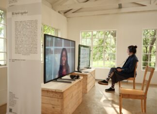 Kunst im Gespräch – Podcastserie Eine Frau sitzt in einem sonnendurchfluteten Raum vor einem Bildschirm.