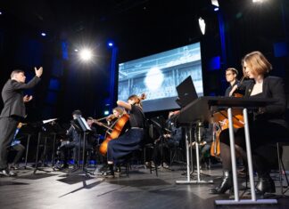 Konzert im Klangtheater der Universität für Musik und darstellende Kunst Wien (mdw) / ASoP Programm Das Bild zeigt das Orchester auf der Bühne, dahinter ein grosser Bildschirm mit einer medialen Darstellung und davor seitlich die Komponistin am Laptop.