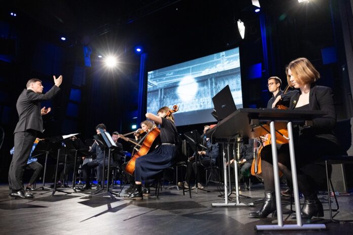 asop-25_(c) stp_web-028-3866 Das Bild zeigt das Orchester auf der Bühne, dahinter ein grosser Bildschirm mit einer medialen Darstellung und davor seitlich die Komponistin am Laptop.
