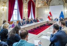 Treffen der EUNIC Gruppe mit Staatssekretär Sepp Schellhorn Am Bild ist ein Sitzungstisch zu sehen, an welchem eine Gruppe von Menschen diskutiert. Der Tisch befindet sich in einem prächtigen Saal mit rotem Teppich und Samtvorhängen.