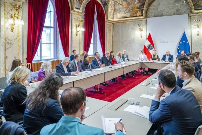 BMEIA Am Bild ist ein Sitzungstisch zu sehen, an welchem eine Gruppe von Menschen diskutiert. Der Tisch befindet sich in einem prächtigen Saal mit rotem Teppich und Samtvorhängen.