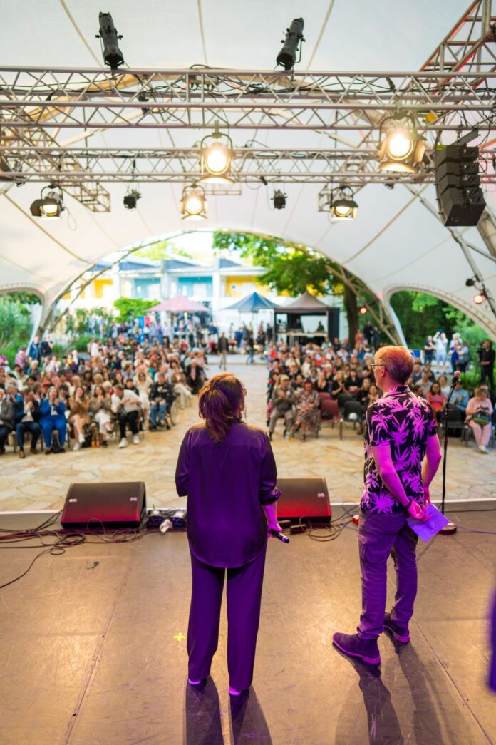 Bild: (c) Jim Kroft Das Bild zeigt die Veranstaltung „Babylon Europa“ auf der Open-Air-Bühne der ufaFabrik Berlin im Jahr 2024. Im Vordergrund stehen zwei Personen auf der Bühne vor einem großen Publikum, von hinten fotografiert: Eine Frau mit dunklen, schulterlangen Haaren in dunkler Kleidung hält ein Mikrofon, daneben steht ein Mann mit hellem Haar, Sonnenbrille und einem Hemd mit pinkem Palmenmuster. Er hält ein gefaltetes Papier in der Hand. Die Bühne ist mit einem weißen Zeltdach überdacht, links und rechts sind zahlreiche Scheinwerfer angebracht. Im Hintergrund sitzt ein vielfältiges Publikum auf Stühlen; einige Menschen stehen, andere applaudieren. Hinter dem Publikum sind bunte Zelte und Gebäude zu sehen – es ist ein sommerlicher Tag. Die Atmosphäre wirkt lebendig und ausgelassen.