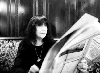 Hommage an Friedericke Mayröcker / Kulturforum Brüssel Das Bild zeigt ein Schwarz-Weiß-Bild der österreichischen Schriftstellerin Friederike Mayröcker, einer älteren Frau mit schulterlangem dunklem Haar. Sie trägt einen schwarzen Pullover und eine lange Halskette, ihre untere Hälfte ist auf dem Bild nicht zu sehen. Sie sitzt auf einer Couch in einem Café und liest eine Zeitung.