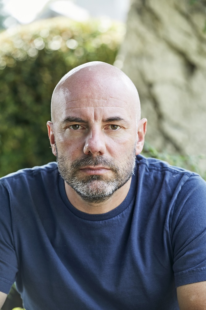 Bild: c) Martin Rauchenwald Das Bild zeigt Daniel Wisser. Er trägt ein blaues T-Shirt. Der Autor hat einen Drei-Tage-Bart und eine Glatze. Er blickt direkt in die Kamera und wirkt nachdenklich.