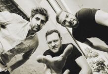 HAEZZ: Jazz aus Österreich / Botschaft Ottawa / Kulturforum Washington / Kulturforum New York Das Bild zeigt drei junge Männer, die sich leicht nach vorne beugen und direkt in die Kamera blicken. Die Aufnahme ist aus einer Froschperspektive gemacht, wodurch die Personen besonders präsent und dynamisch wirken. Einer der Männer in der Mitte streckt eine Hand in Richtung Kamera aus. Alle drei tragen legere Kleidung – zwei in dunklen T-Shirts, einer in einem hellen Hemd. Der Hintergrund besteht aus hellen, leicht verwitterten Wänden und einem schmalen Ausschnitt des Himmels. Das Foto ist in Schwarz-Weiß gehalten und vermittelt eine künstlerisch-urban wirkende Atmosphäre.