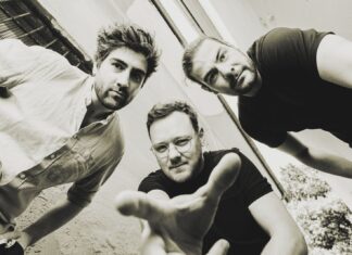 HAEZZ: Jazz aus Österreich / Botschaft Ottawa / Kulturforum Washington / Kulturforum New York Das Bild zeigt drei junge Männer, die sich leicht nach vorne beugen und direkt in die Kamera blicken. Die Aufnahme ist aus einer Froschperspektive gemacht, wodurch die Personen besonders präsent und dynamisch wirken. Einer der Männer in der Mitte streckt eine Hand in Richtung Kamera aus. Alle drei tragen legere Kleidung – zwei in dunklen T-Shirts, einer in einem hellen Hemd. Der Hintergrund besteht aus hellen, leicht verwitterten Wänden und einem schmalen Ausschnitt des Himmels. Das Foto ist in Schwarz-Weiß gehalten und vermittelt eine künstlerisch-urban wirkende Atmosphäre.