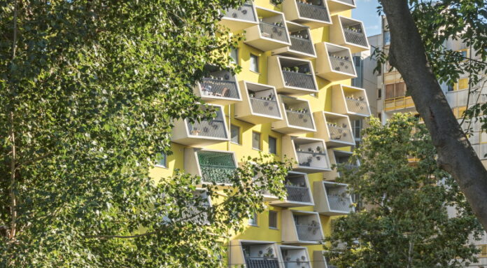 Bild Copyright: © querkraft – hertha hurnaus Das Bild zeigt einen gelben Wohnblock der querkraft architekten mit kleinen Balkonen. Links und rechts ist das Gebäude von Blattwerk umgeben.