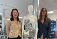 Something for Everyone, Everything for No One / Botschaft Oslo Das Foto wurde bei der Ausstellungseröffnung am 7.5.2025 aufgenommen und zeigt die beiden Künstlerinnen neben einem ihrer Werke. Links steht die US Künstlerin Teak RAMOS. Sie hat schwarze halblange Haare und dunkle Augen. Sie trägt einen schwarzen knielangen weiten Rock und eine beige Jacke, schwarze Strümpfe und Sneaker. Recht steht die österreichische Künstlerin Anna-Sophie BERGER. Sie hat hellbraune lange Haare und helle Augen. Sie trägt eine olivfarbene Hose, eine dunkelblaue Bluse und mit Strasssteinen besetzte schwarze Sandalen. In der Mitte steht ein Second Hand Mannequin das eine der Modeschöpfungen der beiden Künstlerinnen trägt. Ein beiges Leinenkleid, etwa knielang, das mit A4 Ausdrucken verschiedener schwarz-weiß Bilder aus der International Library of Fashion Research benäht ist. Darunter ein Kleeblatt-Autobahnknoten, eine sitzende nähende Frau und ein Schneidertisch.