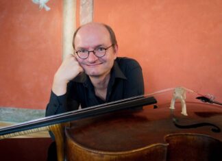 Kunst im Gespräch – Podcastserie Auf dem Bild sitzt Wolfgang Panhofer, vor ihm quer liegend das Cello. Er hat den Kopf auf einer Hand abgestützt und lächelt breit und spitzbübisch in die Kamera. Wolfgang trägt eine Brille und hat eine Halbmondglatze. Er trägt ein schwarzes Hemd, das sich vor dem orangen Hintergrund gut abhebt.