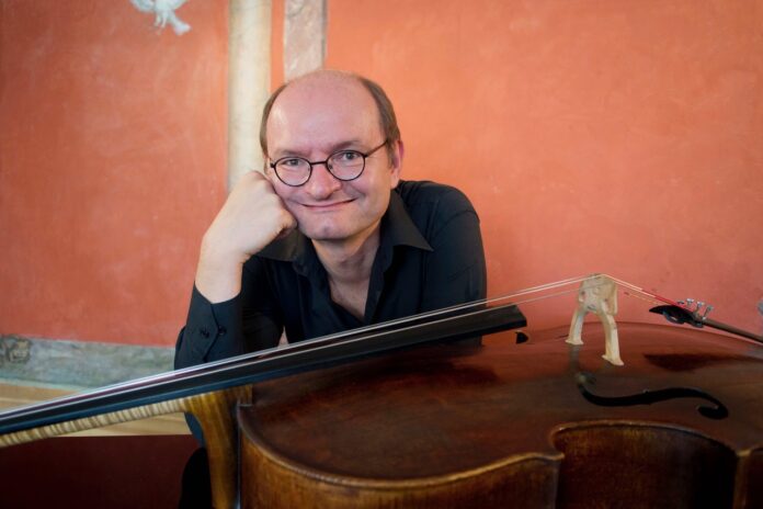 Bild: (c) Christine de Grancy Auf dem Bild sitzt Wolfgang Panhofer, vor ihm quer liegend das Cello. Er hat den Kopf auf einer Hand abgestützt und lächelt breit und spitzbübisch in die Kamera. Wolfgang trägt eine Brille und hat eine Halbmondglatze. Er trägt ein schwarzes Hemd, das sich vor dem orangen Hintergrund gut abhebt.