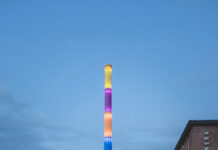 Kunstfestival Begehungen 2025 / Kulturforum Berlin Das Bild zeigt das stillgelegte Heizkraftwerk Chemnitz-Nord in der Dämmerung. Im Zentrum steht ein hoch aufragender Schornstein, der in leuchtenden Regenbogenfarben illuminiert ist. Rechts ist ein Backsteingebäude mit der Aufschrift „HEIZKRAFTWERK“ zu sehen, im Vordergrund ein vorbeifahrendes Auto.