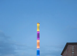 Kunstfestival Begehungen 2025 / Kulturforum Berlin Das Bild zeigt das stillgelegte Heizkraftwerk Chemnitz-Nord in der Dämmerung. Im Zentrum steht ein hoch aufragender Schornstein, der in leuchtenden Regenbogenfarben illuminiert ist. Rechts ist ein Backsteingebäude mit der Aufschrift „HEIZKRAFTWERK“ zu sehen, im Vordergrund ein vorbeifahrendes Auto.