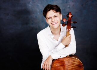 Kirchenkonzert von Jeremias Fliedl / Kulturforum Prag Das Bild zeigt den Cellisten Jeremias Fliedl mit einem weißen Hemd vor einem dunkelgrauen Hintergrund. Er ist gut gelaunt und hält in seiner rechten Hand sein Violoncello am Griffbrett fest, und lehnt sich mit der linken Hand am Korpus des Cellos an.