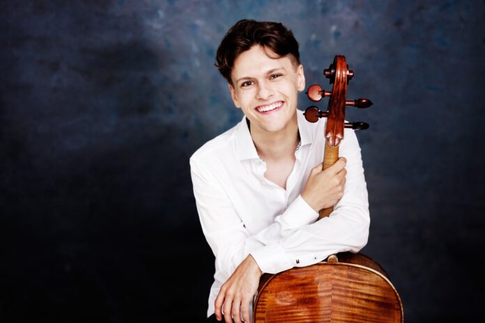 Das Bild zeigt den Cellisten Jeremias Fliedl mit einem weißen Hemd vor einem dunkelgrauen Hintergrund. Er ist gut gelaunt und hält in seiner rechten Hand sein Violoncello am Griffbrett fest, und lehnt sich mit der linken Hand am Korpus des Cellos an.