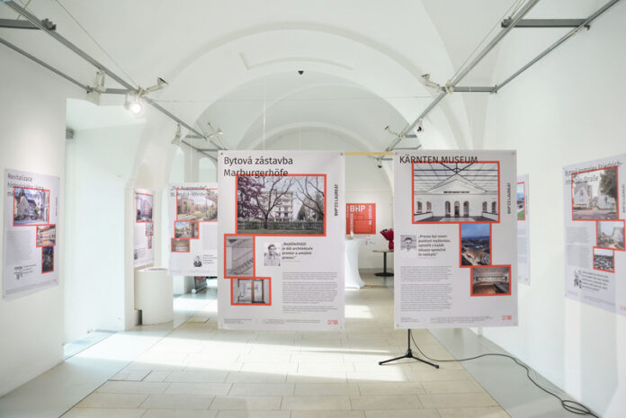 Bild: Tereza Kunderová Das Bild zeigt die Galerie des Österreichischen Kulturforums. Auf beiden Seiten, sowie im Zentrum, kann man die Projekte der Fotoausstellung „BHP Bauherr:innenpreis 2022-2024“ erkennen. Hierbei kann man die Gestaltung der Banner, die jedes einzelne Projekt präsentieren sehen. So gibt es auf der rechten oder linken unteren Seite Informationstext und von oben nach unten Bilder der Projekte, die mit roter Farbe umrahmt sind. Außerdem kann man auf den Bannern Zitate in großem Textformat sehen, wo sich ein Bild des Zitierten befindet. Die zwei Projekte im Mittelpunkt der Kamera sind „Marburgerhöfe“ und „Kärnten Museum“. Die Galerie ist ansonsten menschenleer.