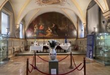 Augarten-Ausstellung in Rom / Botschaft Rom Das Bild zeigt die Sala del Cenacolo mit den Flaggen Italiens und der Europäischen Union im Hintergrund. Ein langer Holztisch in der Mitte des Raumes präsentiert Exponate der Wiener Porzellanmanufaktur Augarten. Entlang der Wände stehen Vitrinen mit weiteren Objekten, begleitet von blauen, informativen Roll-Ups. Im Zentrum des Raumes steht eine große Vase aus der Augarten-Sammlung.