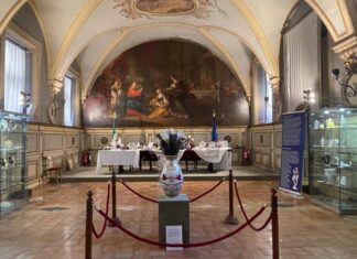 Augarten-Ausstellung in Rom / Botschaft Rom Das Bild zeigt die Sala del Cenacolo mit den Flaggen Italiens und der Europäischen Union im Hintergrund. Ein langer Holztisch in der Mitte des Raumes präsentiert Exponate der Wiener Porzellanmanufaktur Augarten. Entlang der Wände stehen Vitrinen mit weiteren Objekten, begleitet von blauen, informativen Roll-Ups. Im Zentrum des Raumes steht eine große Vase aus der Augarten-Sammlung.