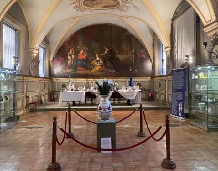 Bild: ÖB Rom – Roberta Koujevetopoulos Das Bild zeigt die Sala del Cenacolo mit den Flaggen Italiens und der Europäischen Union im Hintergrund. Ein langer Holztisch in der Mitte des Raumes präsentiert Exponate der Wiener Porzellanmanufaktur Augarten. Entlang der Wände stehen Vitrinen mit weiteren Objekten, begleitet von blauen, informativen Roll-Ups. Im Zentrum des Raumes steht eine große Vase aus der Augarten-Sammlung.
