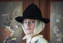 20&20 – A Lens of Her Own / Kulturforum Tel Aviv Das Bild zeigt das Ausstellungsposter: Eine Frau schaut in die Kamera. Sie hat ein weißes Tuch umgebunden und einen Hut auf dem Kopf. Im Hintergrund sind Bilder mit Blumenmuster, auf einer Seite zerrissen, zu sehen.