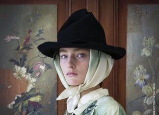 20&20 – A Lens of Her Own / Kulturforum Tel Aviv Das Bild zeigt das Ausstellungsposter: Eine Frau schaut in die Kamera. Sie hat ein weißes Tuch umgebunden und einen Hut auf dem Kopf. Im Hintergrund sind Bilder mit Blumenmuster, auf einer Seite zerrissen, zu sehen.