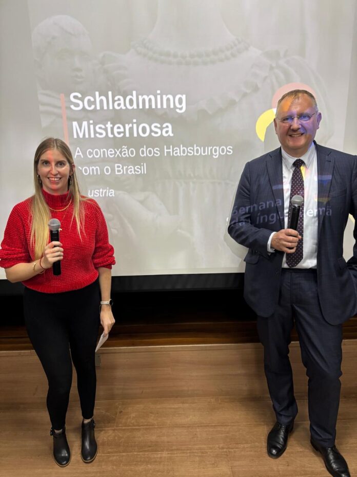 Bild: ÖB Brasília Das Bild zeigt den Filmautor Günter Fuhrmann mit Erstzugeteilter Judith Schildberger vor dem Kinobildschirm mit der Einblendung des Filmplakats von Geheimnisvolles Schladming - Habsburgs Brasilien Connection