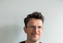 Andreas Jungwirth in Norwegen / Österreichische Botschaft Oslo Der Autor Andreas Jungwirth, Jahrgang 1967, steht mit locker verschränkten Armen und freundlich lächelnd vor hellgrauem Bildhintergrund. Er trägt ein schwarzes kurzärmeliges T-Shirt, eine Brille mit dünnem Rahmen, eine blonde etwas wuschelige Kurzhaarfrisur und einen kurzen grau melierten Bart.