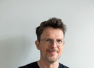 Andreas Jungwirth in Norwegen / Österreichische Botschaft Oslo Der Autor Andreas Jungwirth, Jahrgang 1967, steht mit locker verschränkten Armen und freundlich lächelnd vor hellgrauem Bildhintergrund. Er trägt ein schwarzes kurzärmeliges T-Shirt, eine Brille mit dünnem Rahmen, eine blonde etwas wuschelige Kurzhaarfrisur und einen kurzen grau melierten Bart.