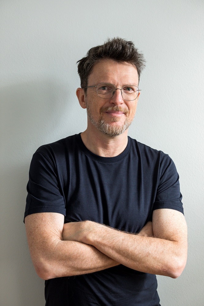 Der Autor Andreas Jungwirth, Jahrgang 1967, steht mit locker verschränkten Armen und freundlich lächelnd vor hellgrauem Bildhintergrund. Er trägt ein schwarzes kurzärmeliges T-Shirt, eine Brille mit dünnem Rahmen, eine blonde etwas wuschelige Kurzhaarfrisur und einen kurzen grau melierten Bart.