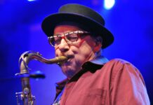 Harry Sokal Groove Unlimited in Brünn / Kulturforum Prag Das Bild zeigt eine Nahaufnahme von Harry Sokal, der konzentriert Saxophon spielt. Er trägt eine schwarz umrandete Brille mit auffälligem Muster in Weiß und Rot sowie einen schwarzen Hut mit runder Krempe. Sein Hemd ist weinrot und leicht gestreift. Das Mundstück des Saxophons liegt fest zwischen seinen Lippen, während er mit geschlossenen Augen ganz in die Musik vertieft scheint. Der Hintergrund ist in tiefem Blaulicht gehalten, typisch für eine Konzertbühne.