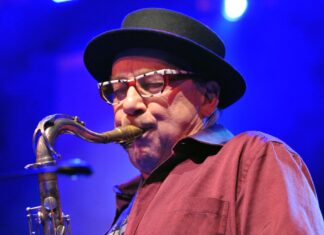 Harry Sokal Groove Unlimited in Brünn / Kulturforum Prag Das Bild zeigt eine Nahaufnahme von Harry Sokal, der konzentriert Saxophon spielt. Er trägt eine schwarz umrandete Brille mit auffälligem Muster in Weiß und Rot sowie einen schwarzen Hut mit runder Krempe. Sein Hemd ist weinrot und leicht gestreift. Das Mundstück des Saxophons liegt fest zwischen seinen Lippen, während er mit geschlossenen Augen ganz in die Musik vertieft scheint. Der Hintergrund ist in tiefem Blaulicht gehalten, typisch für eine Konzertbühne.