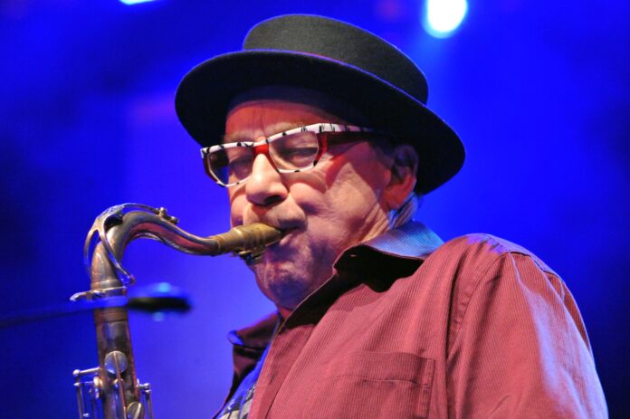 Das Bild zeigt eine Nahaufnahme von Harry Sokal, der konzentriert Saxophon spielt. Er trägt eine schwarz umrandete Brille mit auffälligem Muster in Weiß und Rot sowie einen schwarzen Hut mit runder Krempe. Sein Hemd ist weinrot und leicht gestreift. Das Mundstück des Saxophons liegt fest zwischen seinen Lippen, während er mit geschlossenen Augen ganz in die Musik vertieft scheint. Der Hintergrund ist in tiefem Blaulicht gehalten, typisch für eine Konzertbühne.