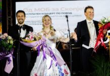 200 Jahre Johann Strauss / Botschaft Skopje Das Bild zeigt einen festlichen Moment am Ende eines Konzerts. Links: Der Bassbariton Ivan Naumovski im eleganten schwarzen Frack mit Fliege, mit einem großen Blumenstrauß in den Armen. In der Mitte: Die Sopranistin Brigitta Simon in einem weißen, festlichen Kleid, sie hält ebenfalls einen prachtvollen Blumenstrauß. Rechts: Der Dirigent Vinzenz Praxmarer in weißer Weste und Frack, ebenfalls mit einem Blumenstrauß in der Hand. Im Hintergrund ist ein großer Bildschirm mit mazedonischem Text zu sehen, auf dem unter anderem auf ein kostenloses Konzert zu Ehren von Johann Strauß hingewiesen wird