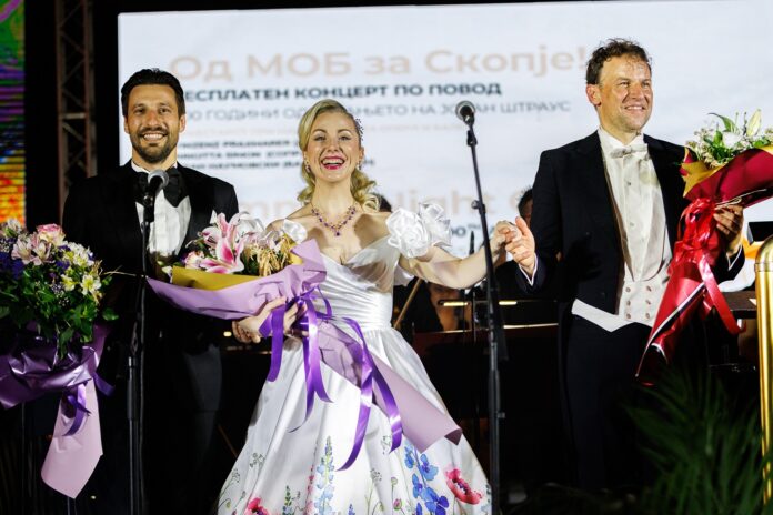Bild: Nationale Oper und Ballett Nordmazedonien Das Bild zeigt einen festlichen Moment am Ende eines Konzerts. Links: Der Bassbariton Ivan Naumovski im eleganten schwarzen Frack mit Fliege, mit einem großen Blumenstrauß in den Armen. In der Mitte: Die Sopranistin Brigitta Simon in einem weißen, festlichen Kleid, sie hält ebenfalls einen prachtvollen Blumenstrauß. Rechts: Der Dirigent Vinzenz Praxmarer in weißer Weste und Frack, ebenfalls mit einem Blumenstrauß in der Hand. Im Hintergrund ist ein großer Bildschirm mit mazedonischem Text zu sehen, auf dem unter anderem auf ein kostenloses Konzert zu Ehren von Johann Strauß hingewiesen wird