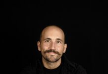 Flying Carpet Children Festival in Mardin / Kulturforum Istanbul Das Bild zeigt einen Mann mit Glatze und Schnurrbart vor schwarzem Hintergrund, der entspannt in die Kamera lächelt.