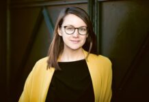 Europäische Literaturnacht / Kulturforum Prag Das Bild zeigt die Schriftstellerin Johanna Seebauer. Sie steht vor einem dunklen, metallischen Hintergrund mit geometrischen Strukturen. Sie hat hellhäutige Haut, schulterlanges braunes Haar, das seitlich gescheitelt ist, und trägt eine schwarze Brille mit runden Gläsern. Sie blickt ruhig und freundlich in die Kamera. Ihre Kleidung besteht aus einem schwarzen Oberteil und einem auffälligen, offenen gelben Mantel oder Blazer.