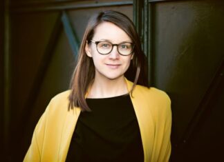 Europäische Literaturnacht / Kulturforum Prag Das Bild zeigt die Schriftstellerin Johanna Seebauer. Sie steht vor einem dunklen, metallischen Hintergrund mit geometrischen Strukturen. Sie hat hellhäutige Haut, schulterlanges braunes Haar, das seitlich gescheitelt ist, und trägt eine schwarze Brille mit runden Gläsern. Sie blickt ruhig und freundlich in die Kamera. Ihre Kleidung besteht aus einem schwarzen Oberteil und einem auffälligen, offenen gelben Mantel oder Blazer.