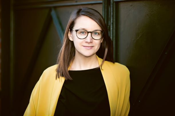 Das Bild zeigt die Schriftstellerin Johanna Seebauer. Sie steht vor einem dunklen, metallischen Hintergrund mit geometrischen Strukturen. Sie hat hellhäutige Haut, schulterlanges braunes Haar, das seitlich gescheitelt ist, und trägt eine schwarze Brille mit runden Gläsern. Sie blickt ruhig und freundlich in die Kamera. Ihre Kleidung besteht aus einem schwarzen Oberteil und einem auffälligen, offenen gelben Mantel oder Blazer.