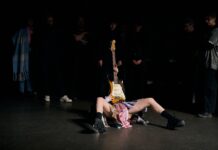 RAGE – Tanzperformance / Kulturforum Tel Aviv Das Bild zeigt die Tänzerin Daphna Horenczyk auf der Bühne liegend mit einer aufgestellten Gitarre.