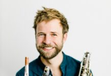 Multitalent Lukas Schiemer / Podcast Auf dem Bild sieht man Lukas Schiemer, sitztend mit einem Saxophon in der Hand. Er trägt eine Jeans und ein blaues Hemd und er lacht in die Kamera. Er hat dunkelblonde Haare und blaue Augen und einen Vollbart.