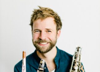 Multitalent Lukas Schiemer / Podcast Auf dem Bild sieht man Lukas Schiemer, sitztend mit einem Saxophon in der Hand. Er trägt eine Jeans und ein blaues Hemd und er lacht in die Kamera. Er hat dunkelblonde Haare und blaue Augen und einen Vollbart.