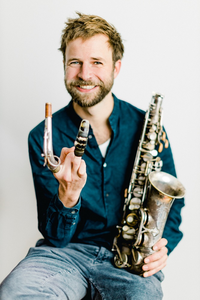 Auf dem Bild sieht man Lukas Schiemer, sitztend mit einem Saxophon in der Hand. Er trägt eine Jeans und ein blaues Hemd und er lacht in die Kamera. Er hat dunkelblonde Haare und blaue Augen und einen Vollbart.