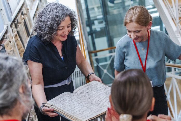BMEIA Es ist eine Frau in einer Bibliothek zu sehen, die vor sich eine grosse alte Urkunde aufgeschlagen hat und darauf blickt.
