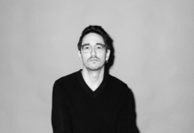 Lesung Marko Dinić / Kulturforum Berlin Das Bild zeigt eine schwarz-weiß Porträtfotografie des Autors Marko Dinić, der auf einem Sessel sitzt. Er hat die Beine überschlagen, die Hände liegen auf den Knien und die Finger sind verschränkt. Er schaut mit ernstem Blick direkt in die Kamera. Der Hintergrund ist einfärbig. Der Autor trägt eine Brille, einen schwarzen Pullover und eine hellere Hose.