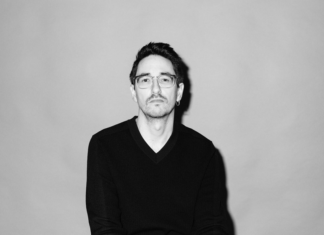 Lesung Marko Dinić / Kulturforum Berlin Das Bild zeigt eine schwarz-weiß Porträtfotografie des Autors Marko Dinić, der auf einem Sessel sitzt. Er hat die Beine überschlagen, die Hände liegen auf den Knien und die Finger sind verschränkt. Er schaut mit ernstem Blick direkt in die Kamera. Der Hintergrund ist einfärbig. Der Autor trägt eine Brille, einen schwarzen Pullover und eine hellere Hose.