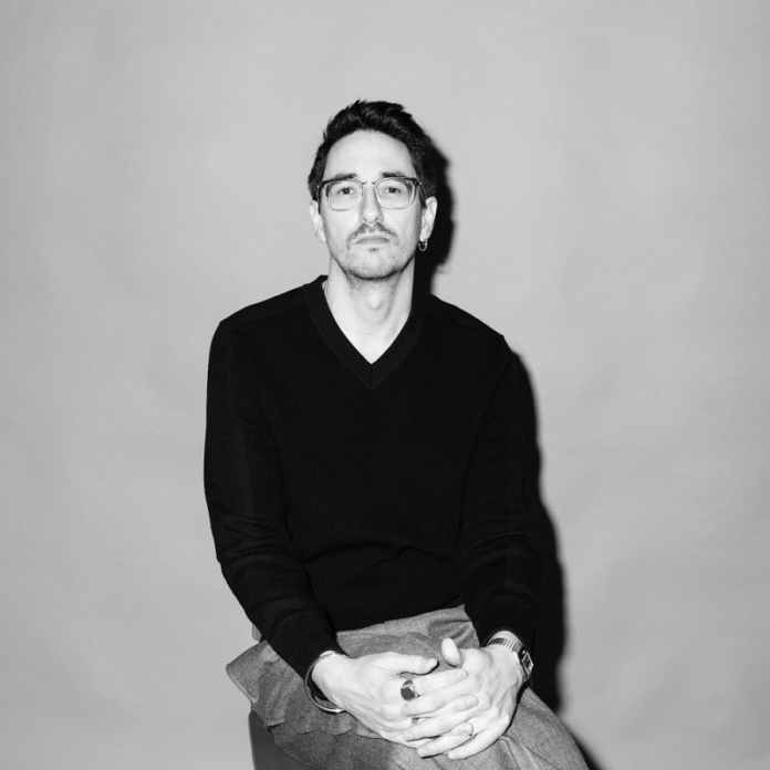 Das Bild zeigt eine schwarz-weiß Porträtfotografie des Autors Marko Dinić, der auf einem Sessel sitzt. Er hat die Beine überschlagen, die Hände liegen auf den Knien und die Finger sind verschränkt. Er schaut mit ernstem Blick direkt in die Kamera. Der Hintergrund ist einfärbig. Der Autor trägt eine Brille, einen schwarzen Pullover und eine hellere Hose.