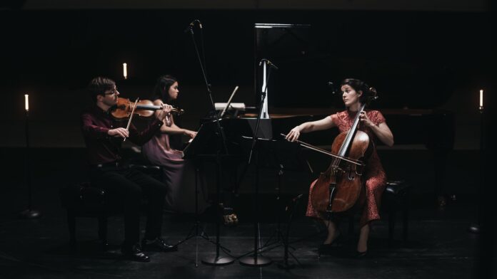 Das Bild zeigt die drei Musiker des accio piano trio, die in einem dunklen Raum spielen. Von links nach rechts sieht man einen Mann, der Geige spielt, eine Frau, die weiter hinten Klavier spielt, und dann, gegenüber dem Mann sitzend, eine Frau, die Cello spielt.