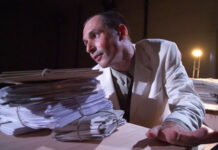 Telephone Conversations with Jandl  / Kulturforum London Das Bild zeigt den Dramaturgen und Darsteller Stephan Bruckmeier, wie er inmitten von Stapeln Papieren sitzt, die linke Hand am Schreibtisch abgegestützt, den Mund halb geöffnet, sein Blick auf ein vermeintliches Publikum gerichtet. Er trägt einen weißen Blazer, im Hintergrund scheint ein Bühnenlicht.