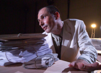 Telephone Conversations with Jandl / Kulturforum London Das Bild zeigt den Dramaturgen und Darsteller Stephan Bruckmeier, wie er inmitten von Stapeln Papieren sitzt, die linke Hand am Schreibtisch abgegestützt, den Mund halb geöffnet, sein Blick auf ein vermeintliches Publikum gerichtet. Er trägt einen weißen Blazer, im Hintergrund scheint ein Bühnenlicht.