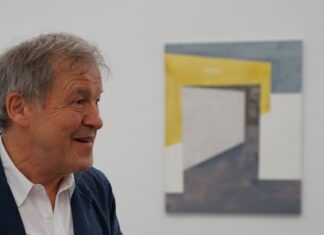 Closing: two dots one line / Kulturforum New York Ein älterer Mann mit grauen Haaren und heller Haut, bekleidet mit einem weißen Hemd und dunkelblauem Sakko, spricht gestikulierend. Er lächelt leicht und schaut nach links. Im Hintergrund hängt ein abstraktes Kunstwerk an der weißen Wand, bestehend aus geometrischen Formen in Gelb, Weiß und Blau. Im Vordergrund ist unscharf ein Teil einer weiteren Person zu sehen.